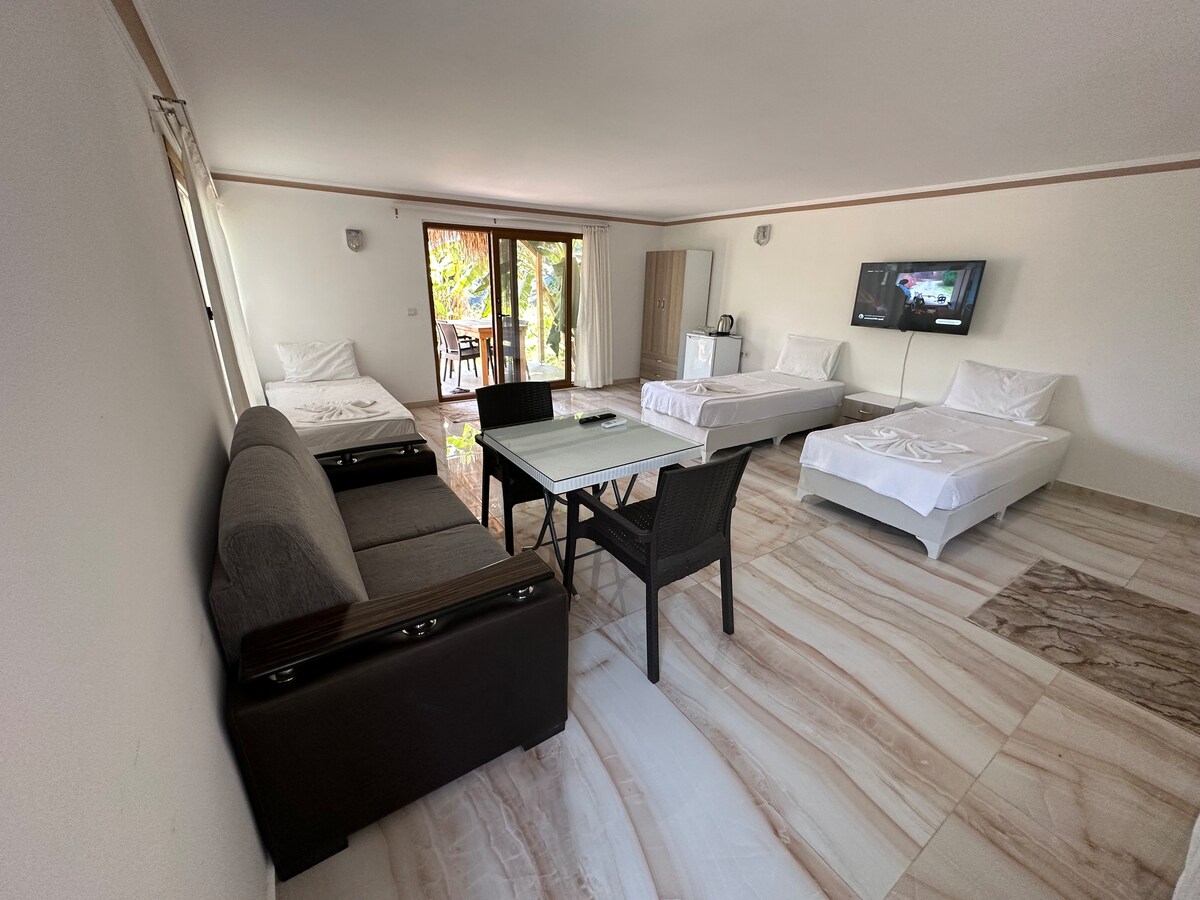 An open living space features two spacious beds and a comfortable sofa. A glass table is positioned in the center, surrounded by dark wicker chairs. Natural light flows in through sliding glass doors leading to an outdoor area, with light-colored tiles enhancing the bright ambiance.