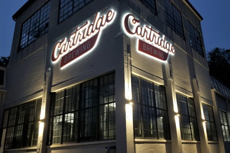 The exterior of Cartridge Brewing is illuminated at night, highlighting its large windows and industrial-style architecture. The prominent signage above the entrance showcases the name 'Cartridge Brewing,' enhancing the inviting atmosphere of this historic building.