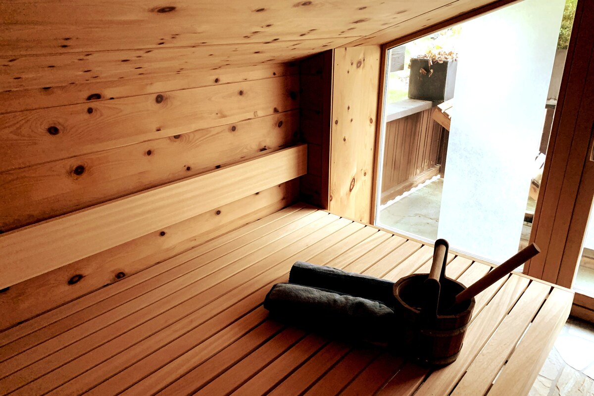 A wooden sauna environment is depicted, featuring smooth wooden benches and a large window that brings in natural light. A sauna bucket and a set of rolled towels are arranged neatly on the bench, adding to the serene ambiance.