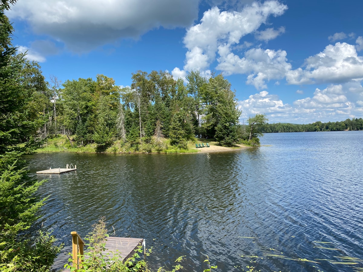 A serene lake scene is depicted with clear blue waters reflecting a sky dotted with fluffy clouds. Lush green trees border the shoreline, and a small sandy beach is visible in the distance. A wooden dock leads toward the water, enhancing the tranquil atmosphere.