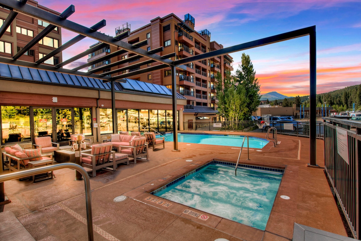 An outdoor area features a heated pool and hot tub, surrounded by a spacious patio with comfortable seating arrangements. Lush trees provide a natural backdrop, while the setting sun casts warm hues across the sky, creating a serene atmosphere.