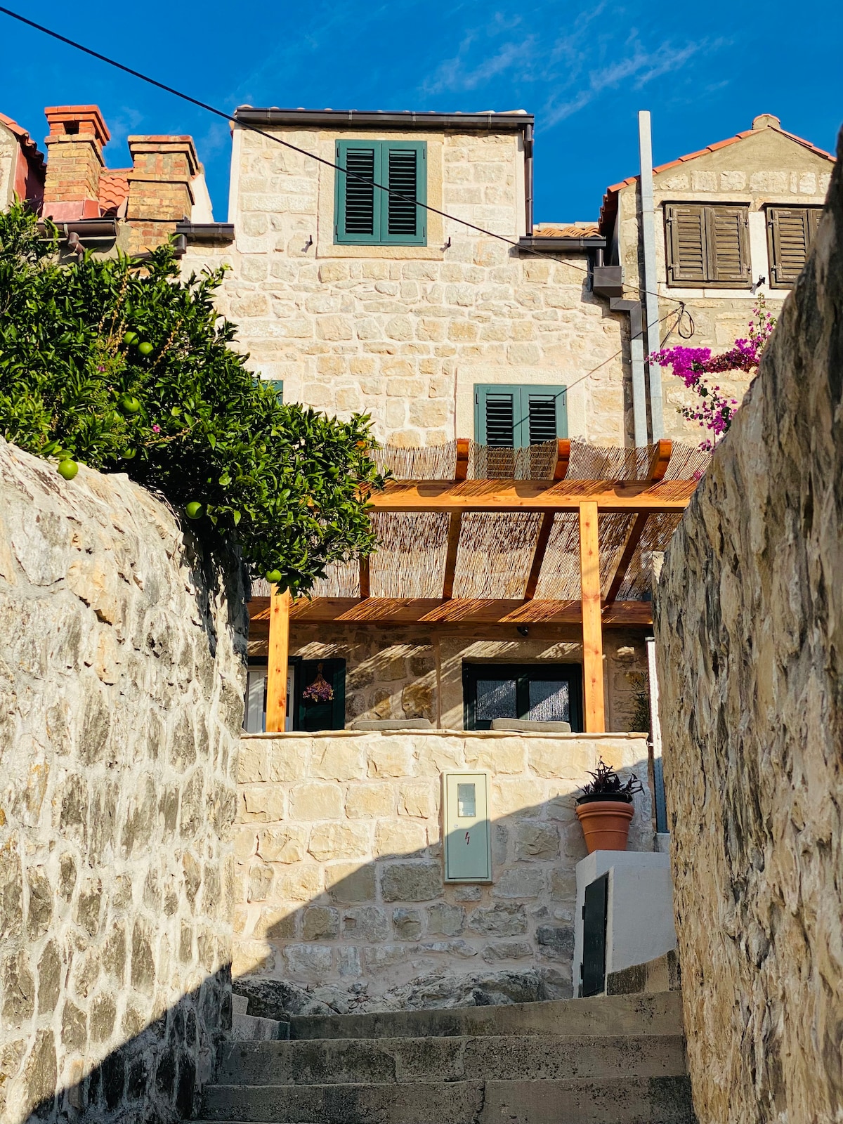 A charming stone villa is framed by vibrant greenery and climbing flowers. The warm-toned facade features green shutters and a wooden pergola, creating a welcoming entrance. Stairs lead up to the home, highlighting the rustic structure within the picturesque old city setting.