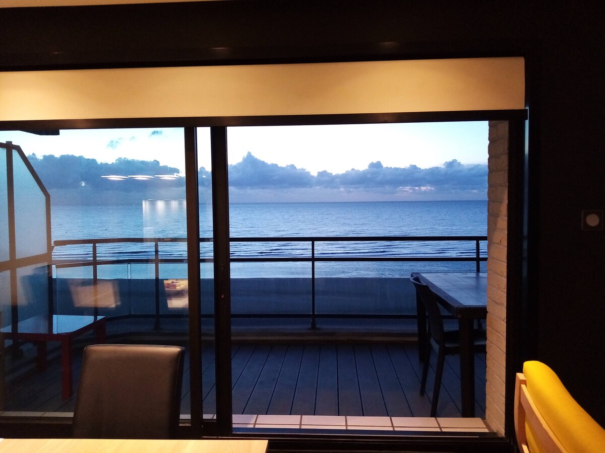 A large glass window offers a view of the tranquil sea and sky, with soft clouds reflecting early morning light. A spacious balcony with outdoor furniture is visible, enhancing the connection with the serene coastal landscape.