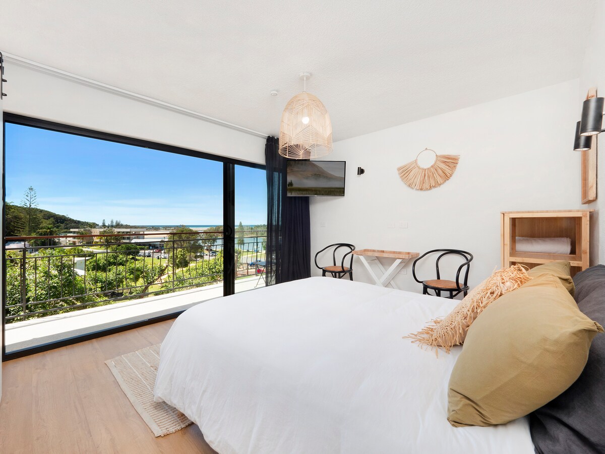 A spacious bedroom features a queen bed with neutral-toned bedding, complemented by decorative pillows. A large sliding glass door provides stunning views of the surrounding landscape. A wooden table and two black chairs are set near the window, while a small shelf holds additional bedding.
