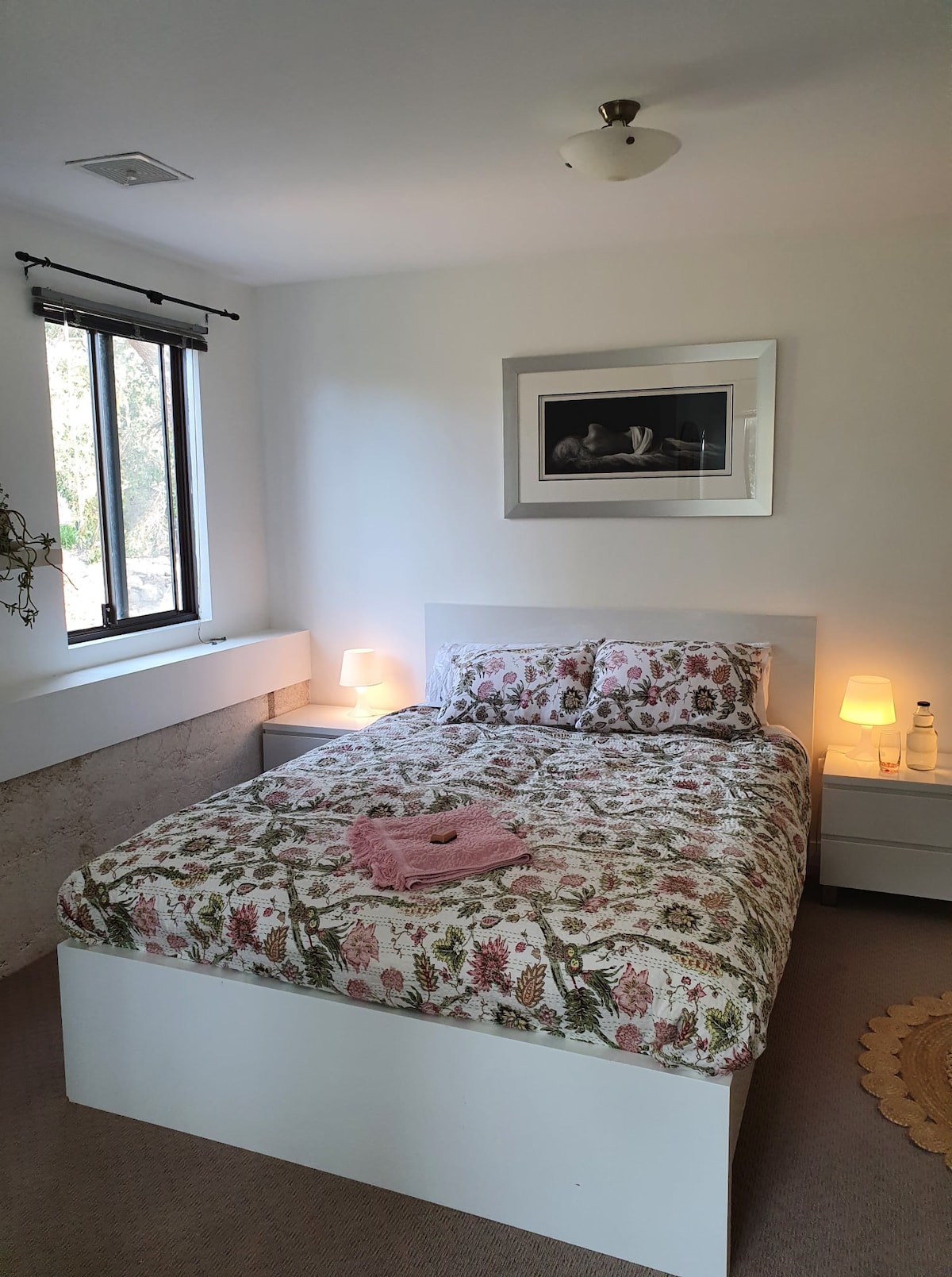 A cozy bedroom features a spacious bed with floral-patterned bedding. Soft lighting from bedside lamps enhances the serene atmosphere. A framed artwork hangs above the bed, while a window allows natural light to fill the room, creating a bright and inviting space.