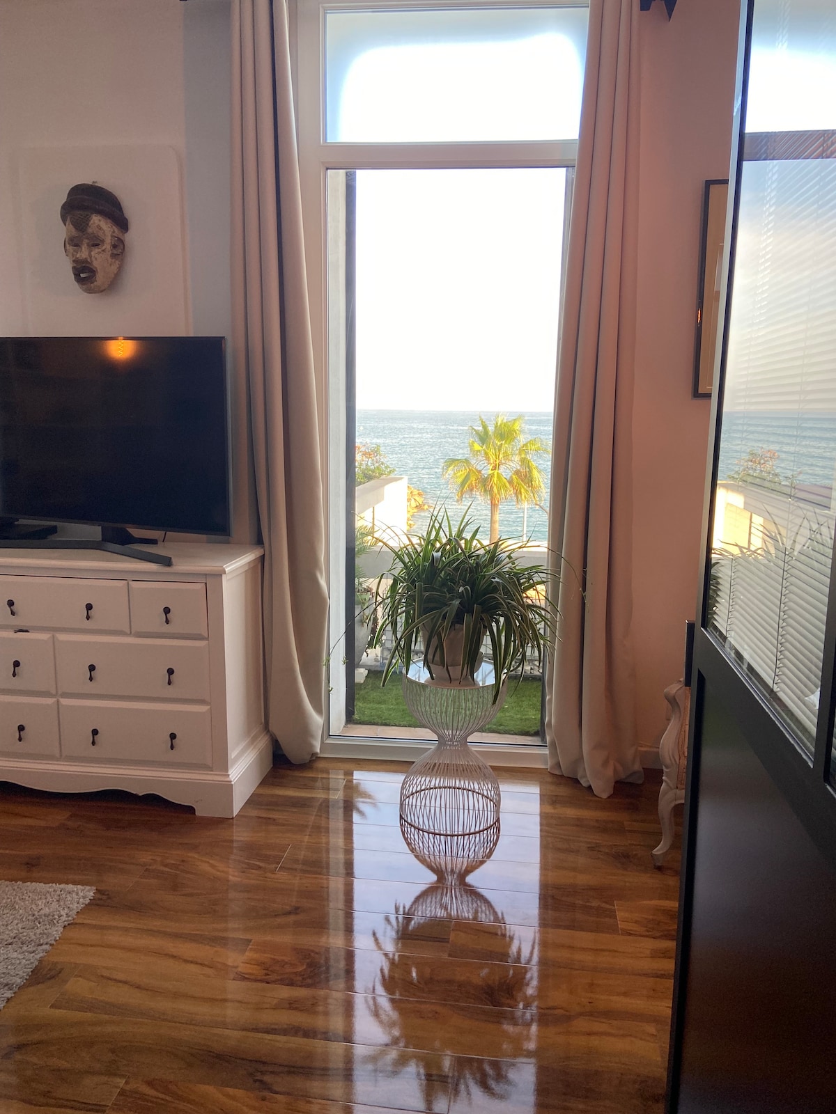 A bright living area features a television on a white dresser, with light streaming through a large window offering a view of palm trees and the ocean. The polished wooden floor reflects the natural light, creating an airy atmosphere.