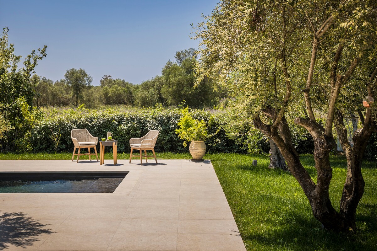 An inviting outdoor area features two rattan chairs positioned beside a small table near the pool. Lush green grass surrounds the space, and olive trees provide shade, creating a serene atmosphere suitable for relaxation.