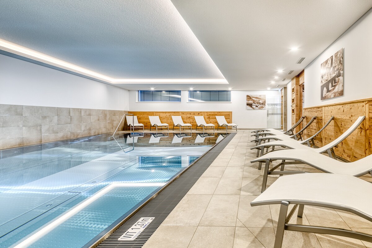 An indoor pool area is featured, with a clear blue swimming pool bordered by light-colored tiles. Adjacent to the pool, several lounge chairs are arranged for relaxation, complemented by warm wooden paneling on one wall, providing a serene atmosphere.