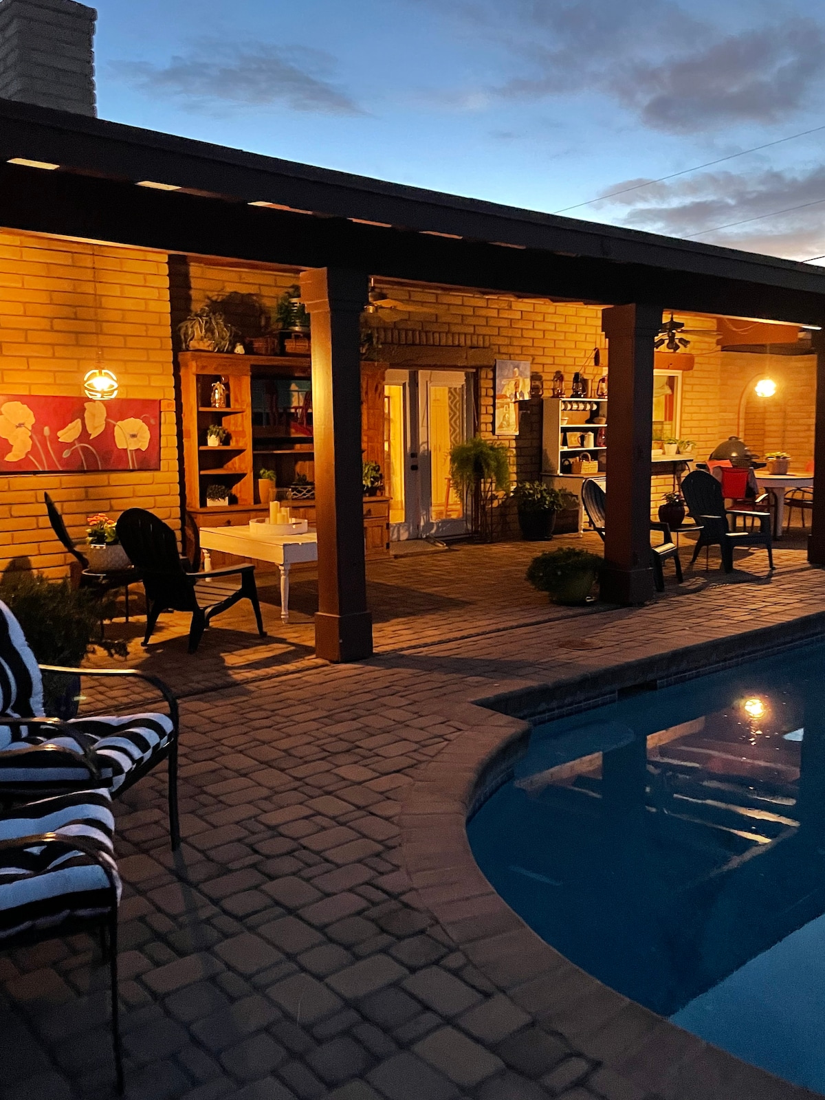 An outdoor patio area is illuminated softly by warm lights, featuring a brick wall with colorful artwork. A seasonal pool reflects nearby greenery, while dark wooden beams provide structure above. Various seating options are arranged around a dining table, creating a cozy atmosphere for gatherings.