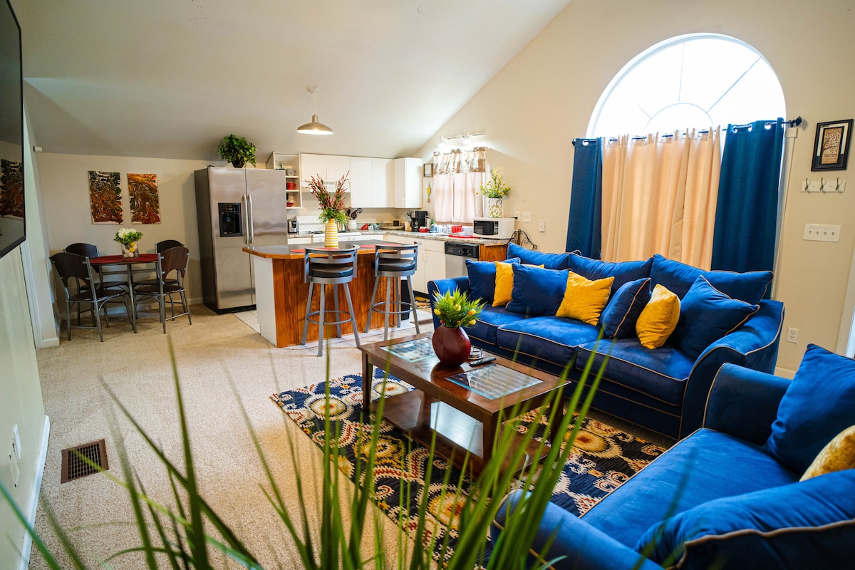 A spacious living area is furnished with a large blue sofa adorned with colorful cushions. A decorative rug anchors the seating space, while a dining area with metal chairs is visible in the background. An arched window allows natural light to brighten the open layout.