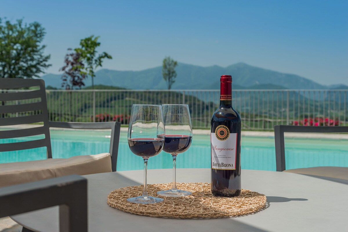 Two glasses of red wine are positioned beside a bottle on a round, woven placemat atop a table. In the background, a turquoise salt-water pool reflects the clear blue sky and rolling green hills, creating a serene atmosphere.