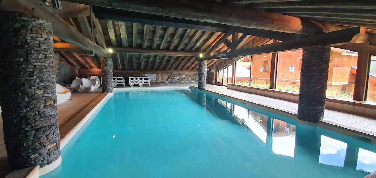 An indoor heated pool features bright turquoise water, surrounded by rustic wooden beams and stone pillars. Large windows allow natural light to fill the space, offering views of the outdoor landscape. Lounge chairs are positioned along the poolside for relaxation.