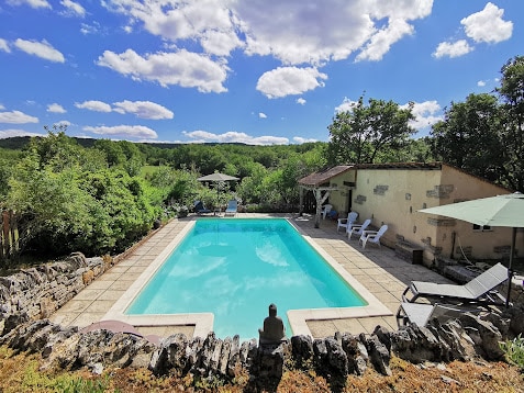 A private swimming pool is surrounded by natural greenery, with sun loungers positioned nearby. A shaded area with umbrellas provides relief from the sun. The pool features clear water, reflecting the sky and clouds above, creating a serene outdoor environment.