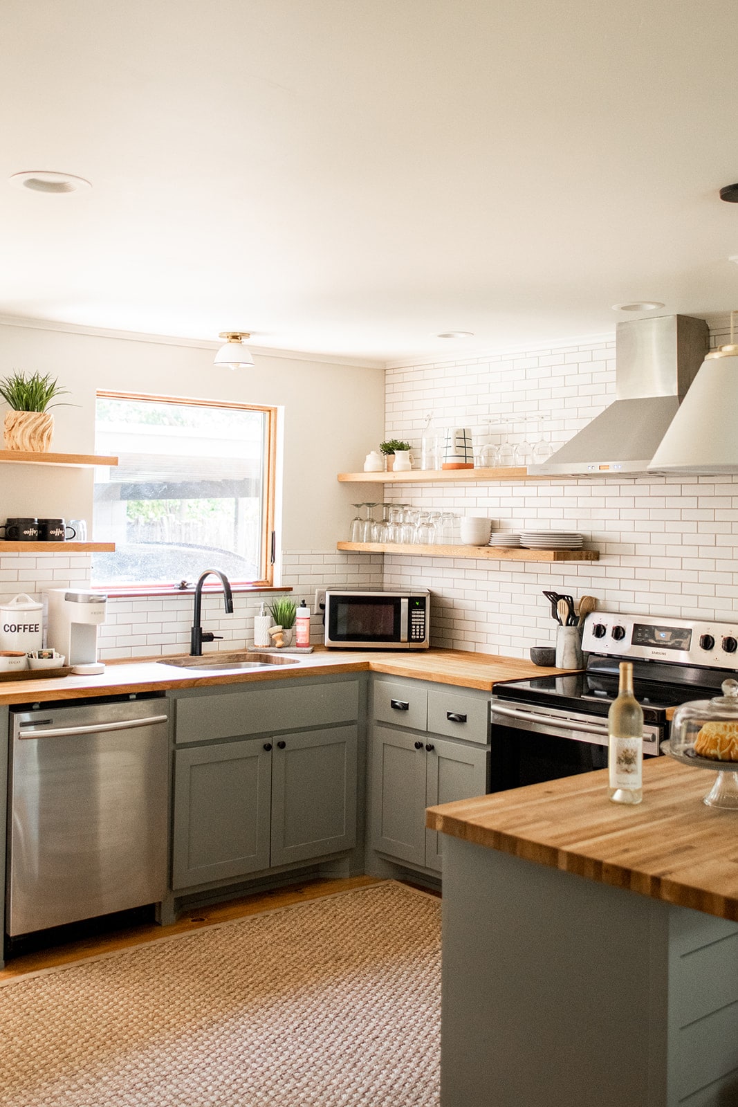 A modern kitchen is designed with light grey cabinetry and wooden countertops. Stainless steel appliances, including an oven and microwave, are present. Open shelving displays dishware and plants, while a window allows natural light to fill the space.
