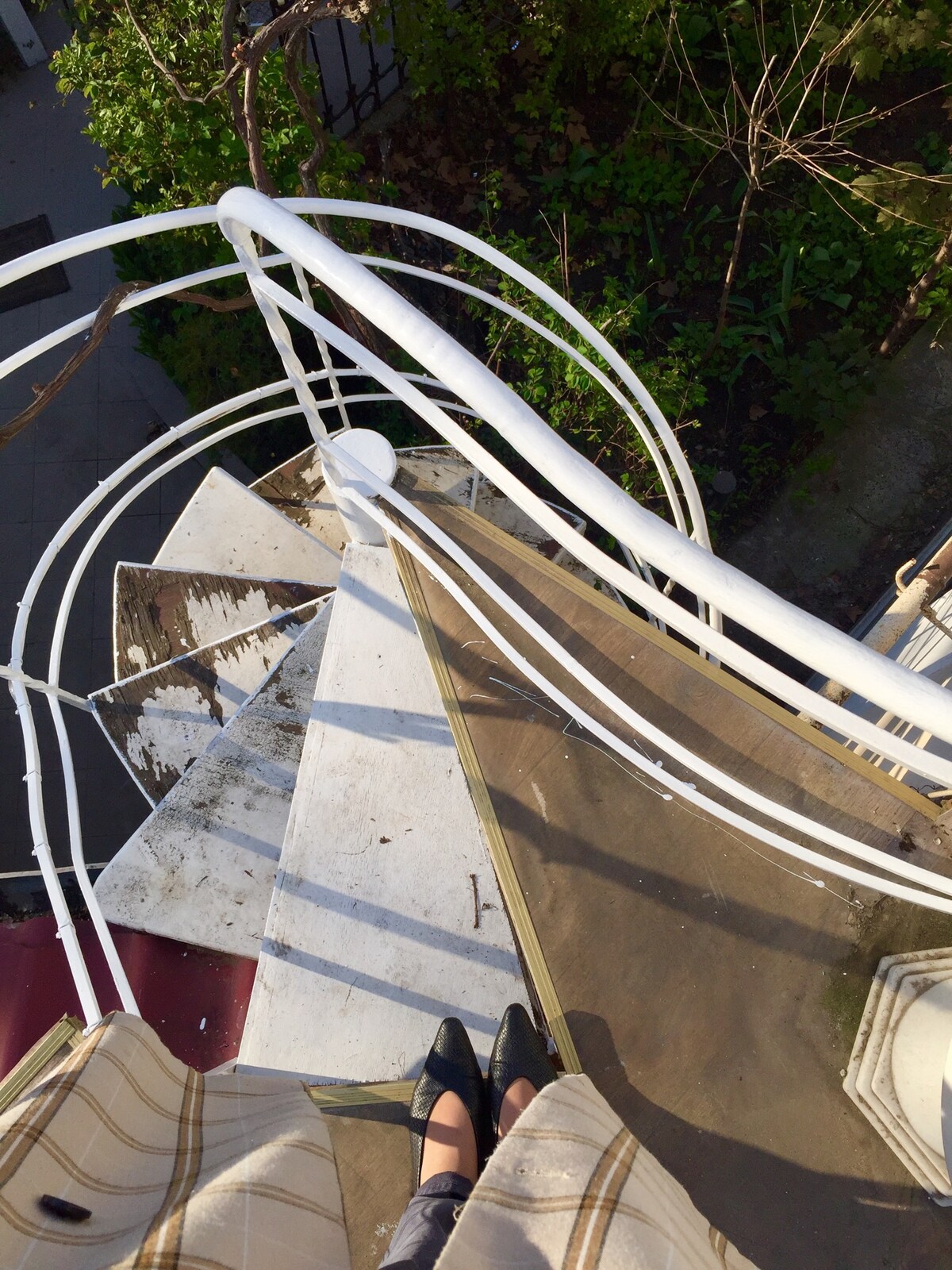 A spiral staircase is featured from an elevated perspective, showcasing white railings. The stairs are adorned with weathered steps, and patches of greenery are visible below. A soft light highlights the textures of the staircase, creating an immersive view of the outdoor space.