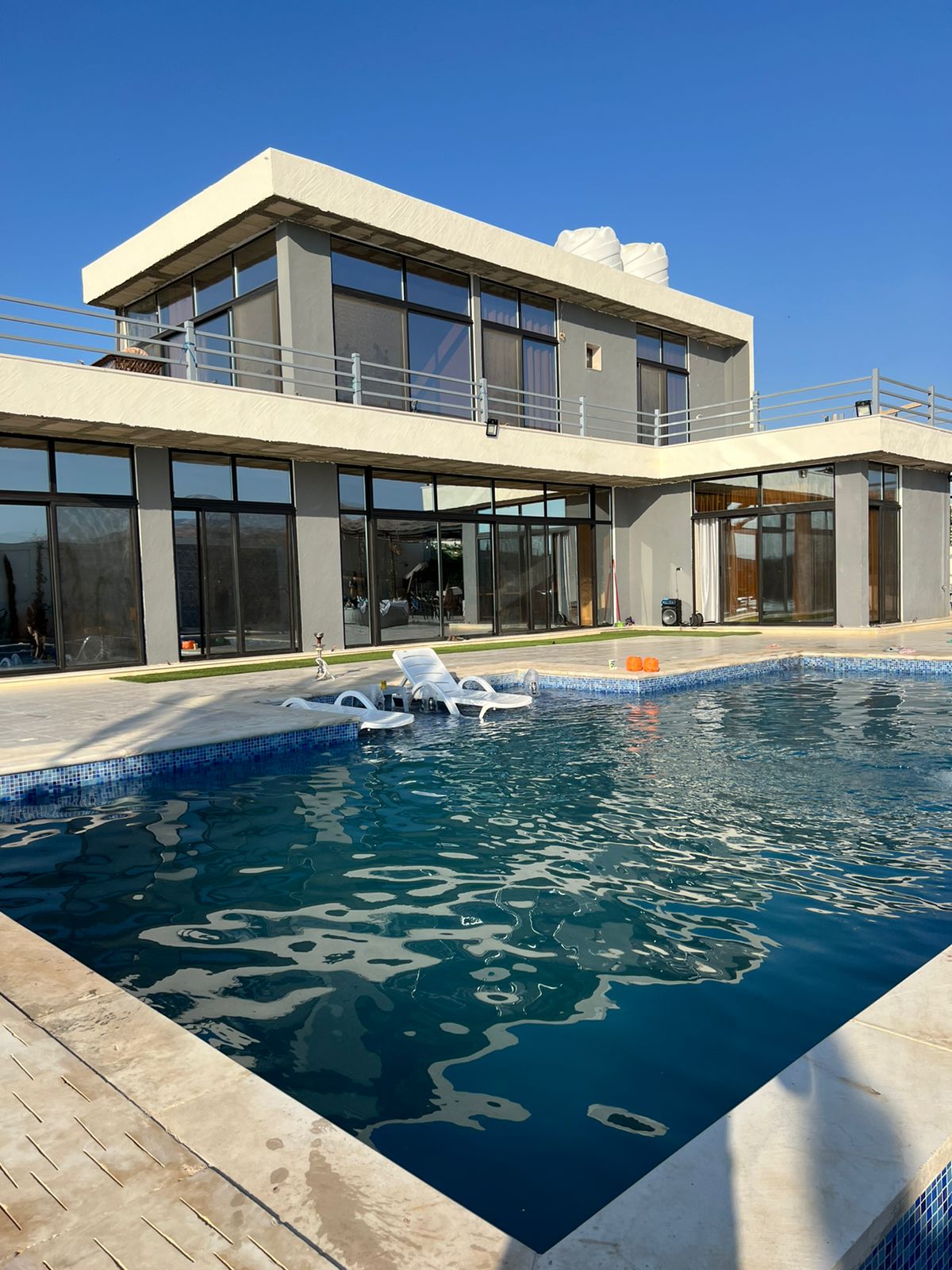 An outdoor swimming pool is framed by a modern villa featuring large glass doors that provide views of the surrounding area. The pool area includes lounge chairs and is set against a clear blue sky, enhancing the open and airy atmosphere.