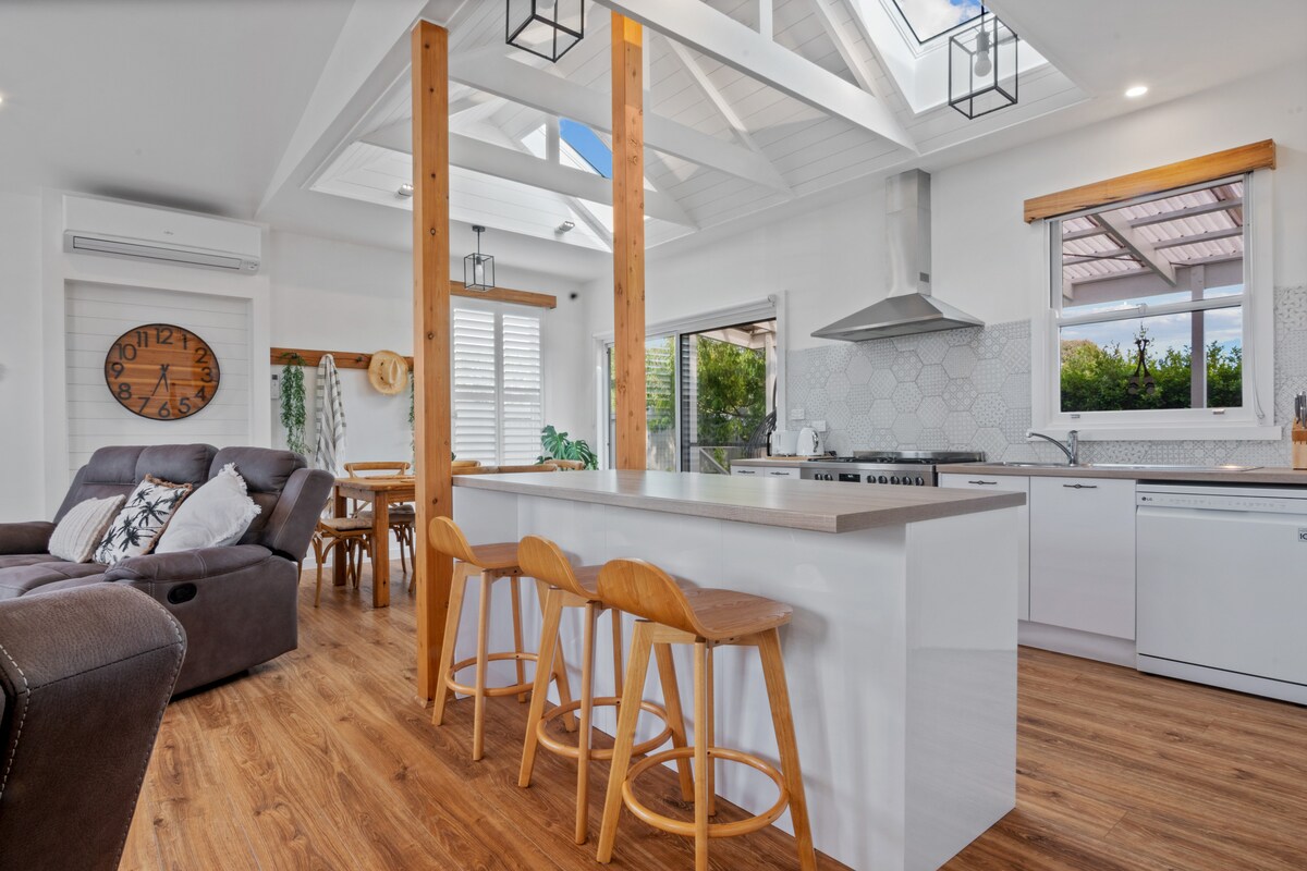 A modern kitchen is showcased, featuring white cabinetry and a large island with seating for three. Skylights allow natural light to fill the space, and a dining area is visible in the background. The room is accented by wood beams, enhancing the airy atmosphere.