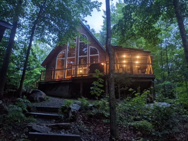 An A-frame cabin is nestled among lush greenery, illuminated softly by outdoor lighting. The large windows reflect warmth and offer a glimpse of the cozy interior. Stone steps lead up to the entrance, surrounded by trees and natural foliage.