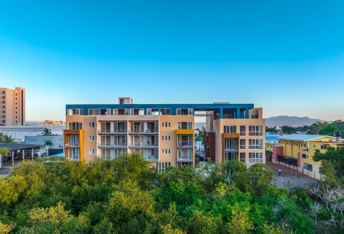The building showcases a modern architectural style, featuring multiple balconies and large windows. It is surrounded by greenery, providing a natural contrast to its contemporary design. The clear blue sky serves as a backdrop, highlighting the tranquility of the setting.