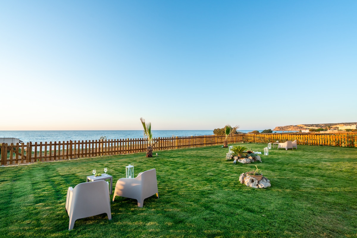 A spacious garden area is presented, featuring a lush green lawn with well-trimmed grass. Two stylish lounge chairs are positioned facing the ocean, inviting relaxation. A stone fire pit is visible, surrounded by decorative stones, while palm trees add a tropical touch in the background.