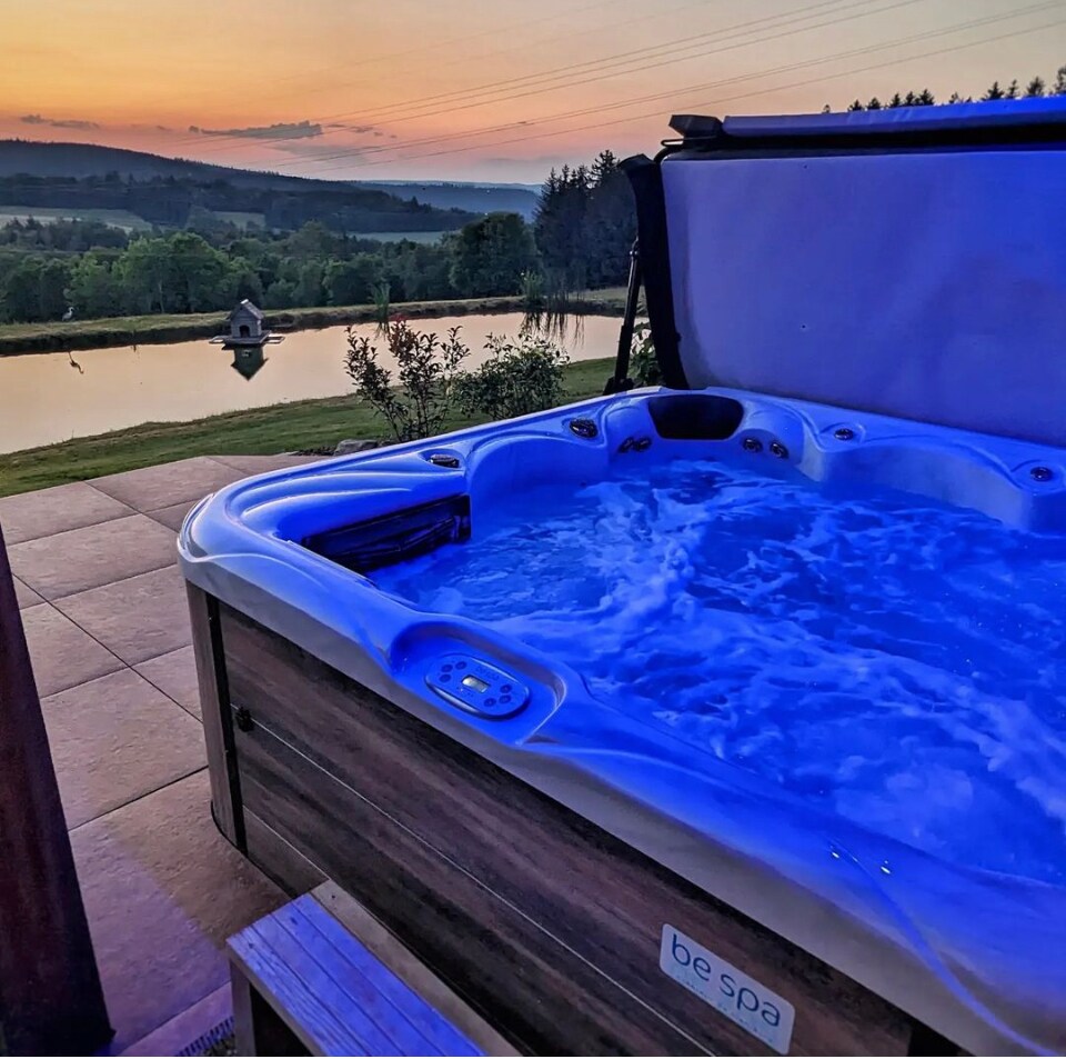 A hot tub with smooth edges is positioned on a stone patio, featuring soft blue lighting. In the background, a serene body of water reflects the colors of a sunset, surrounded by lush greenery and distant hills.