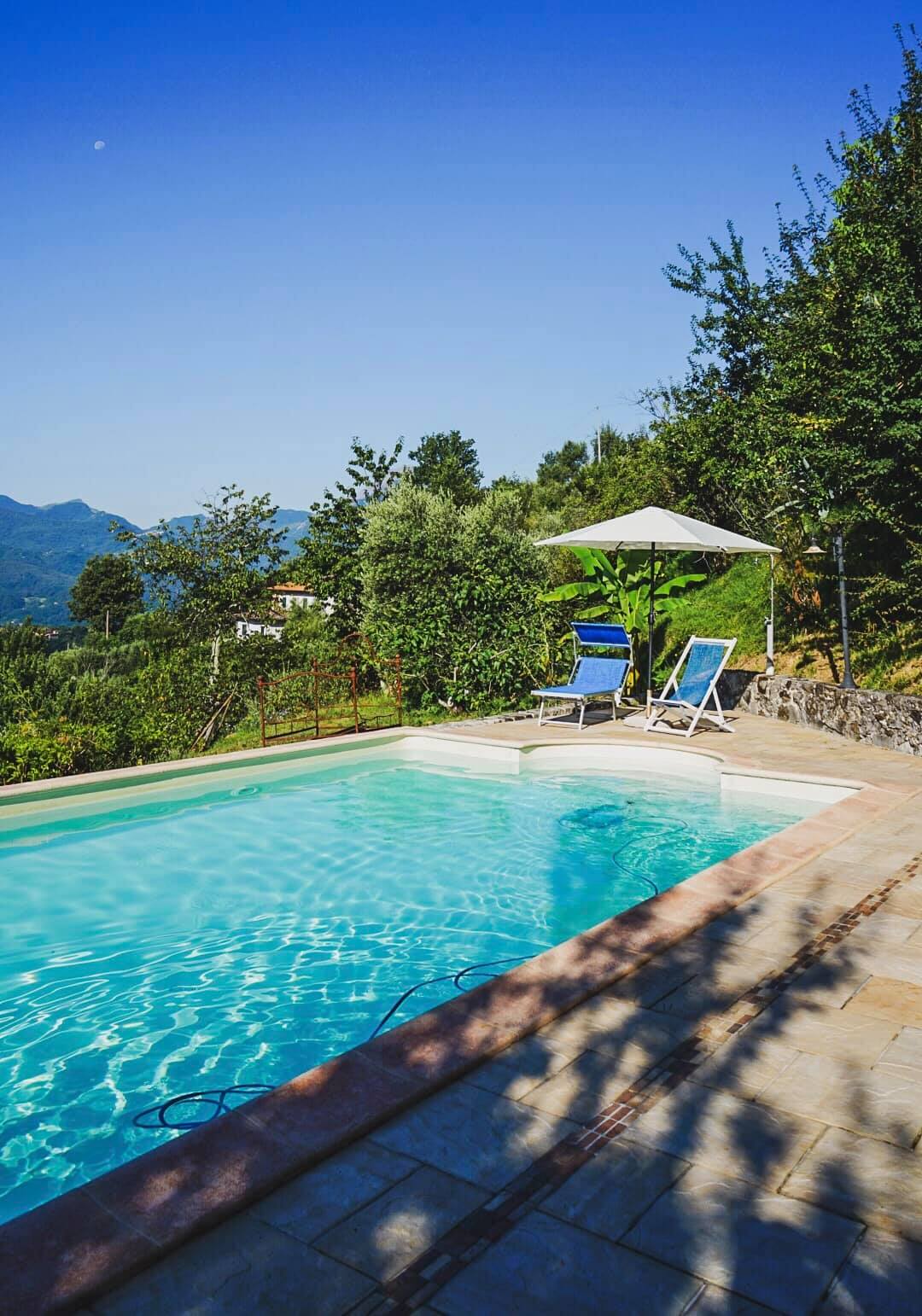 A clear swimming pool is surrounded by lush greenery, with lounge chairs positioned under a shade umbrella. The pool area offers scenic views of the surrounding hills under a bright blue sky, creating a peaceful spot for relaxation.