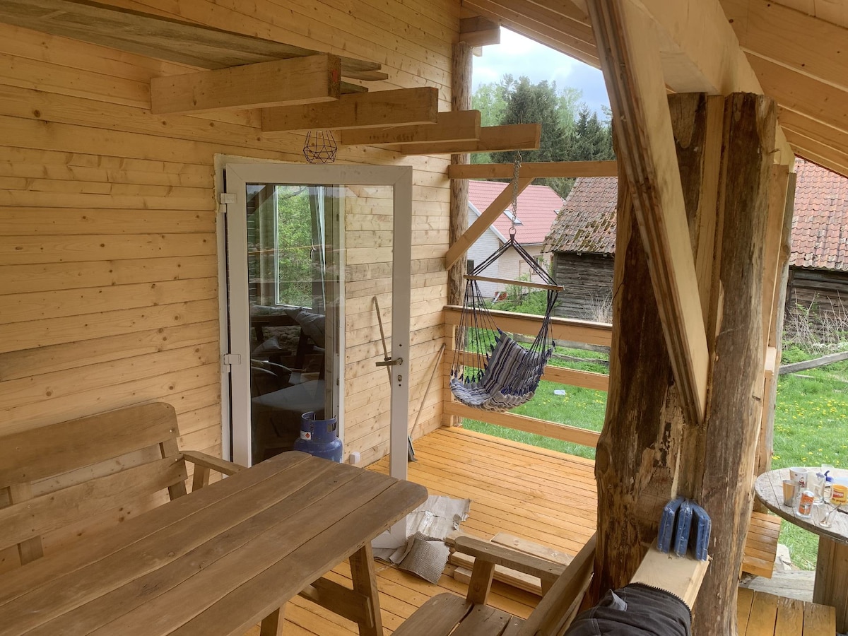 A wooden porch features a sturdy dining table and chairs, complemented by a hammock for relaxation. Large glass doors provide access to the interior, while the surrounding nature is visible through the wooden beams of the structure.