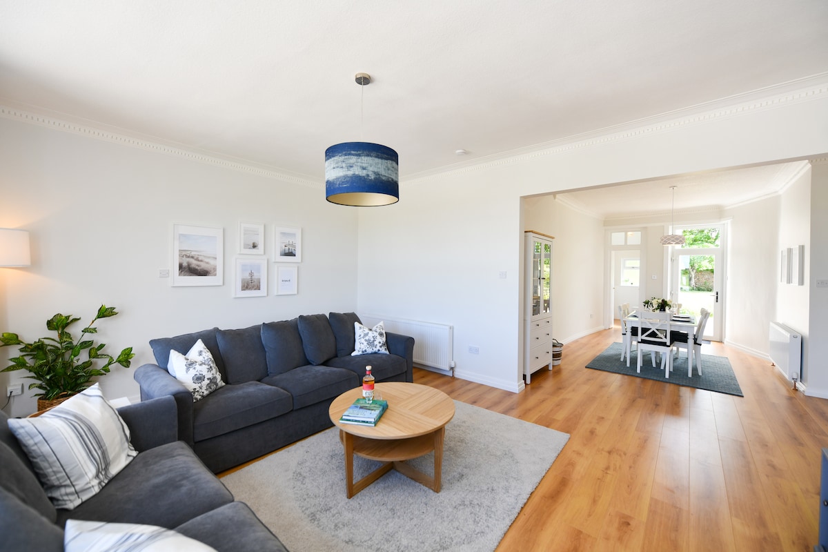 A spacious living area is furnished with a large gray sectional sofa, complemented by a coffee table. The room features hardwood flooring and natural light from large windows. The dining space is visible in the background, adorned with a dining table and chairs.