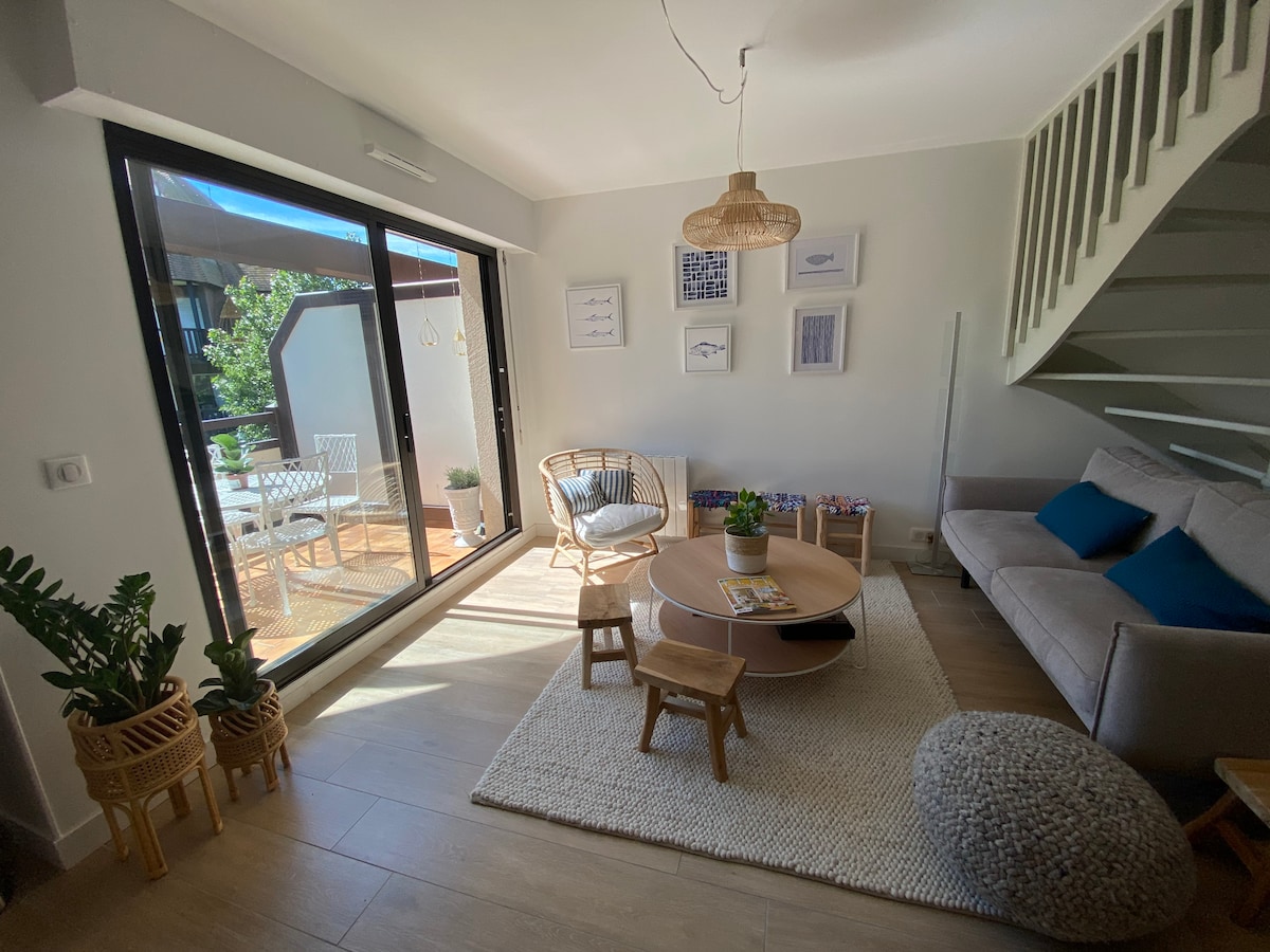 A bright living area features large sliding glass doors opening to a sunny terrace. A comfortable sofa rests against the wall, complemented by a round coffee table and two small stools. Natural light illuminates the space, highlighting decorative wall art and a textured rug.