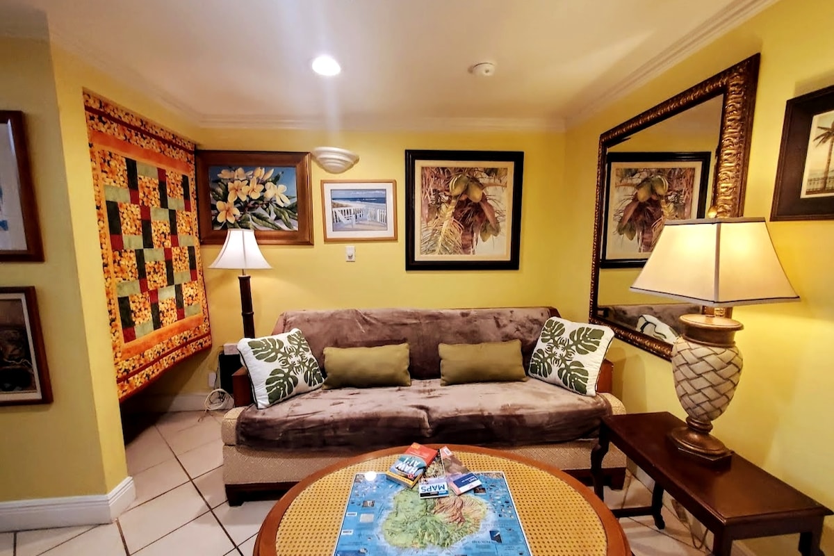 A welcoming living area is presented, featuring a comfortable sofa adorned with tropical-patterned cushions. A round coffee table holds a map of Kauai, while vibrant Hawaiian art decorates the walls. Two lamps provide soft lighting, enhancing the cozy atmosphere.