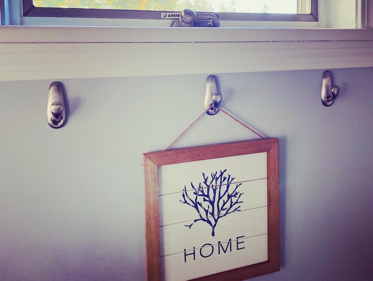 A wall displays three metallic hooks, each designed to hold items. Below them, a wooden sign with a tree design and the word 'HOME' is prominently featured. Natural light enters through the nearby window, illuminating the serene blue walls.