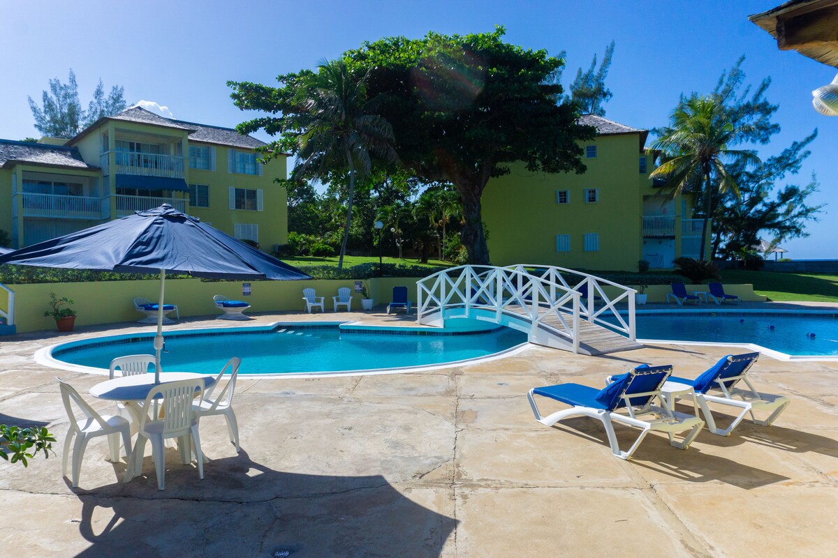A serene outdoor pool area features a circular pool surrounded by a sun deck with blue lounge chairs. A shaded table with white chairs offers a place to relax. Lush greenery and palm trees enhance the tropical atmosphere, with buildings visible in the background.