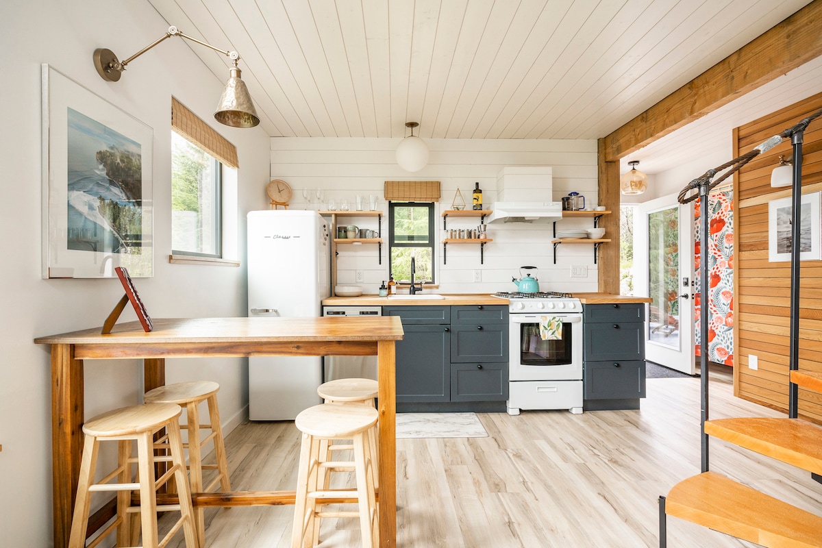 A modern kitchen is presented with light wood ceilings and white walls. Open shelving displays dishes and decorative items. A farmhouse sink and stove are visible in a dark cabinetry setup. A small dining table with three wooden stools completes the inviting space.