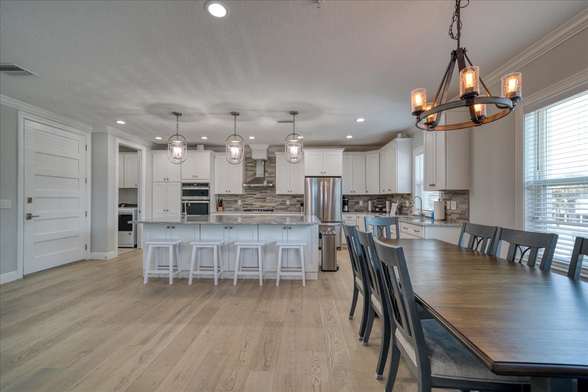 A spacious kitchen and dining area features a large wooden table with seating for eight, complemented by pendant lighting. A central island with barstool seating for four enhances functionality. Stainless steel appliances and neutral cabinetry are visible, creating a modern, inviting space.