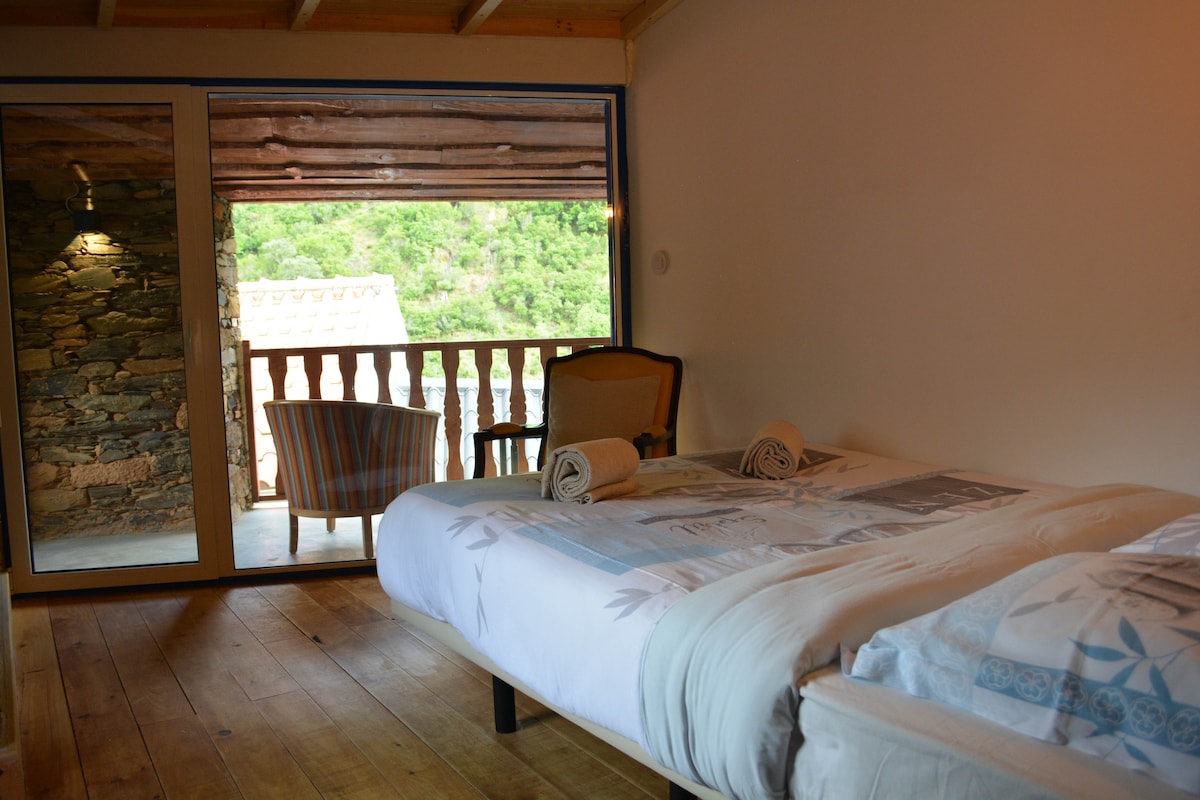 A bright sleeping area features two spacious beds adorned with light bedding. Rolled towels are placed neatly on each bed. A chair and small table are positioned by a glass door leading to a balcony, showcasing views of the surrounding natural landscape.