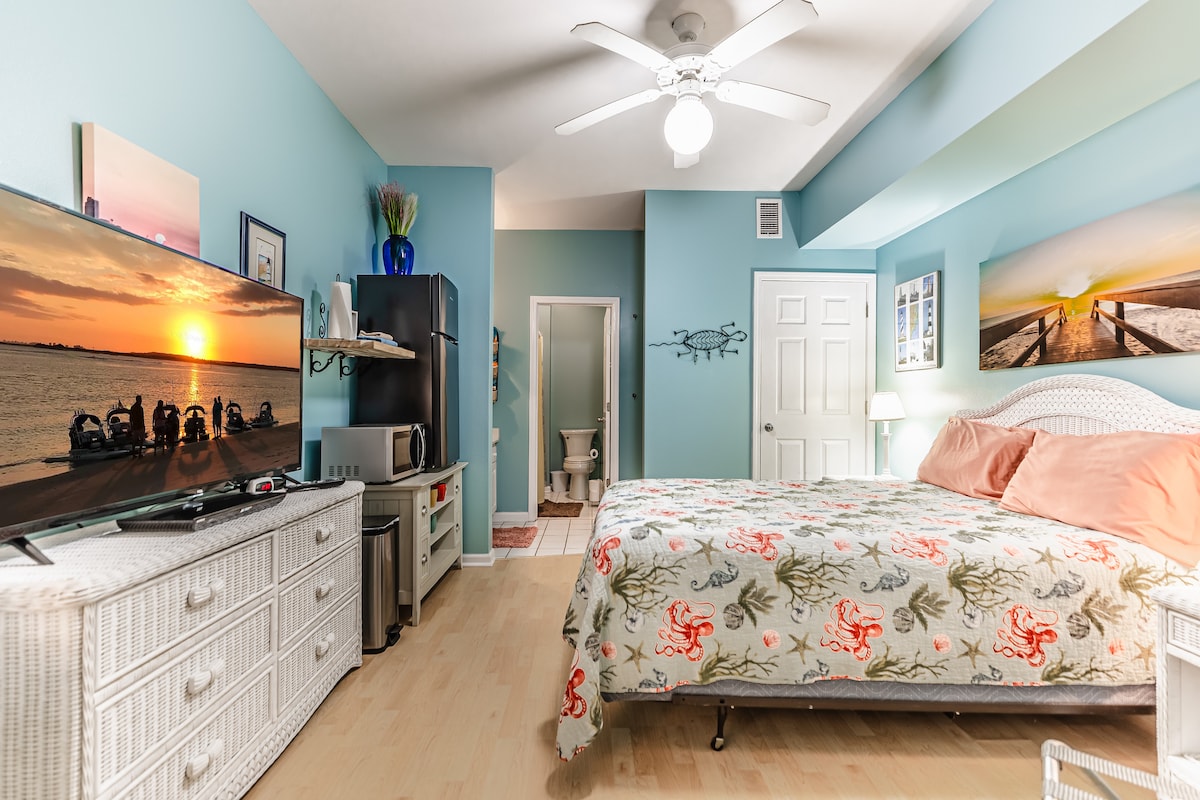 A small, private guestroom features a spacious bed adorned with floral bedding. A large television is mounted on a light-colored dresser. Soft blue walls complement the decor. A bathroom door is visible in the background, and natural light illuminates the room from a nearby window.