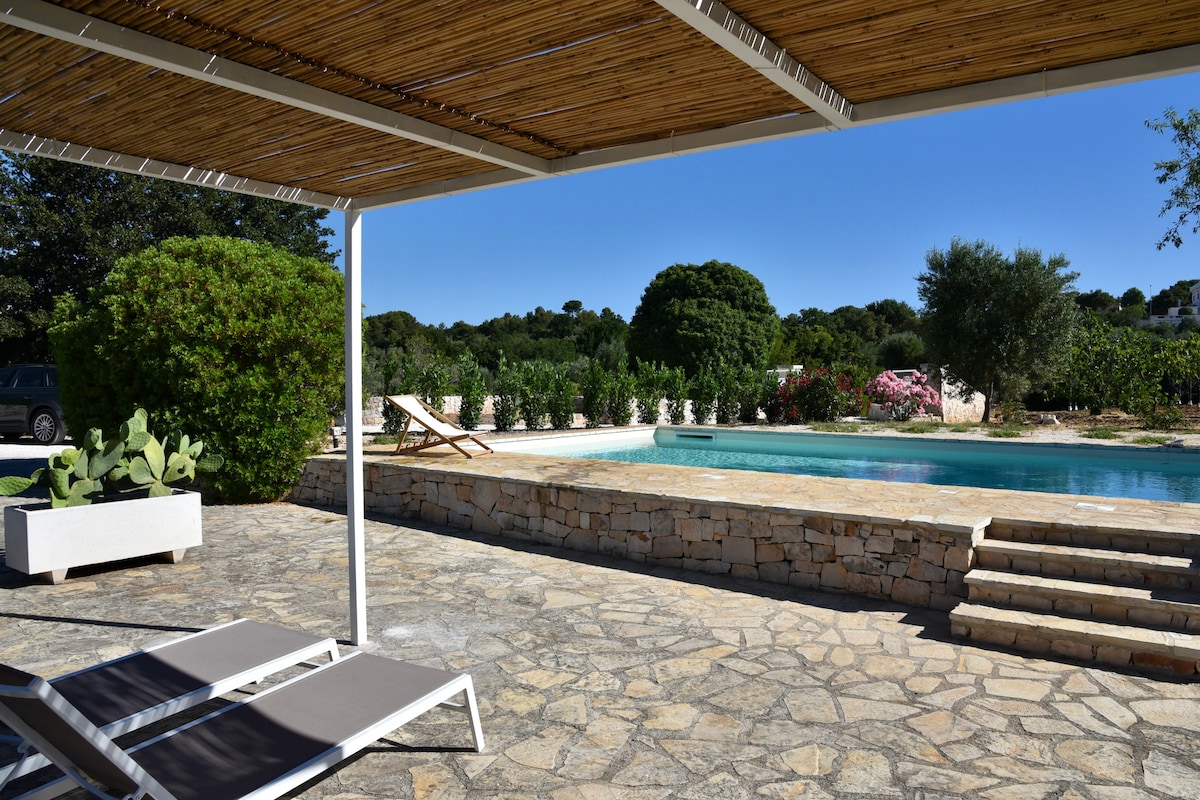 An outdoor patio area is presented with stone flooring, featuring two lounge chairs and a large, shaded seating space. A private pool is visible in the background, framed by lush greenery and blooming flowers, creating a serene outdoor retreat.