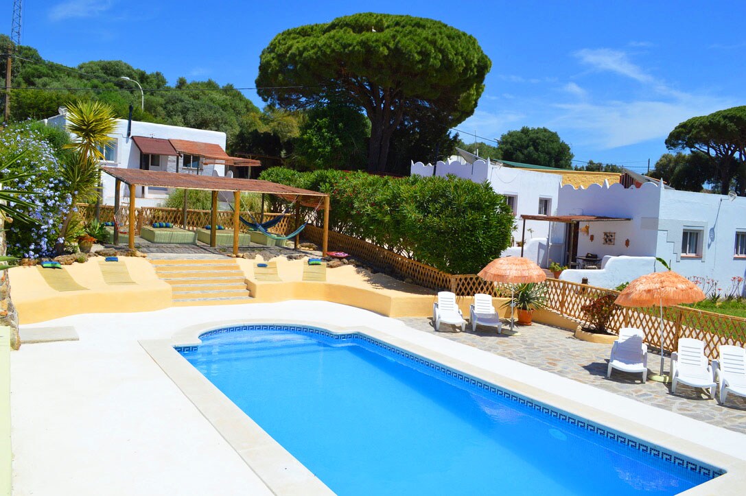 A clear view of the inviting swimming pool surrounded by landscaped gardens. Sun loungers are arranged around the pool, while a shaded seating area is visible nearby. Multi-level terraces lead up from the pool, framed by lush greenery and blue skies.