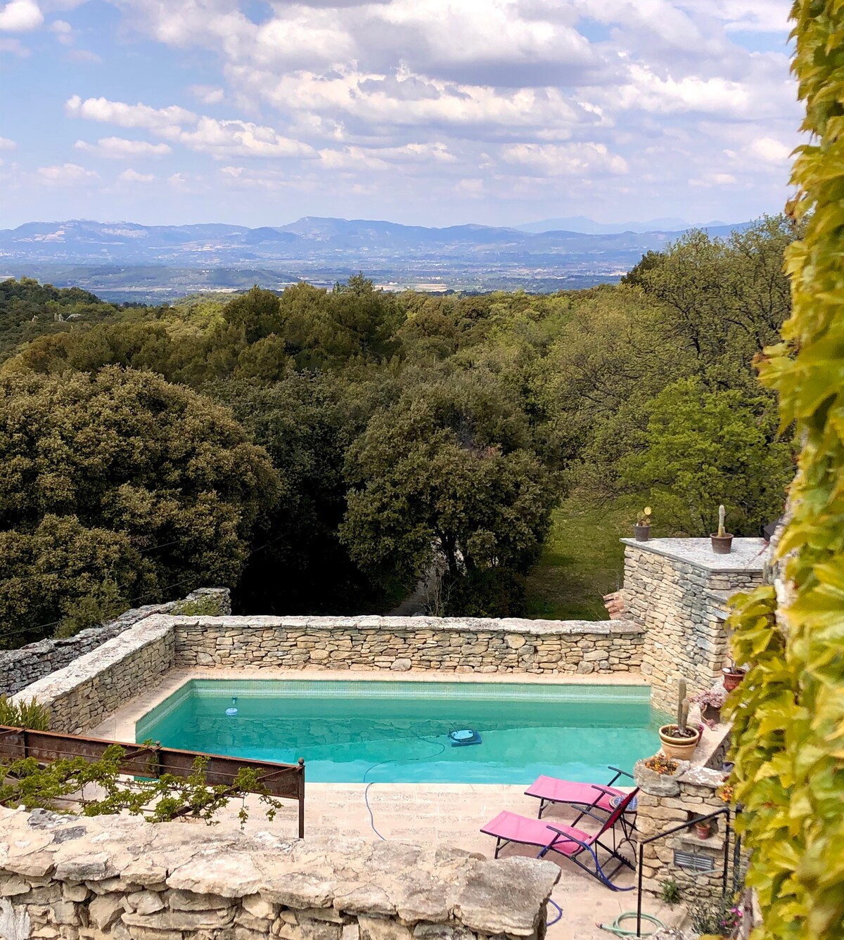 The outdoor area features a private pool surrounded by natural stone walls and lush greenery. Two purple lounge chairs are positioned nearby, offering a serene vantage point overlooking the expansive mountainous landscape under a partly cloudy sky.