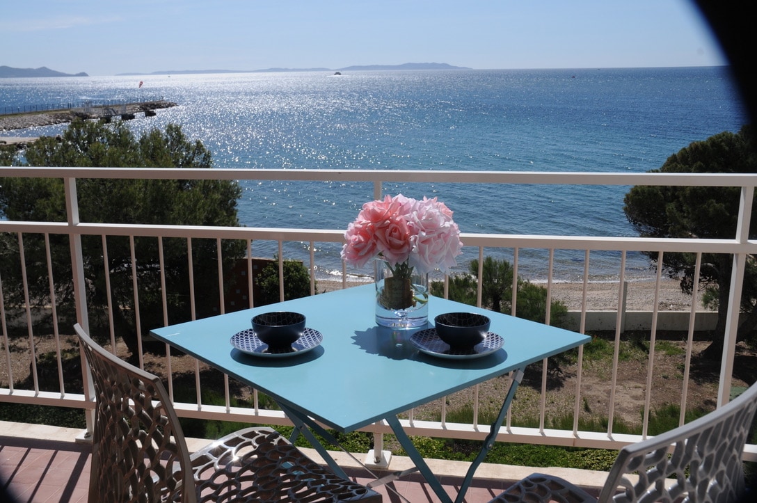 A terrace with a turquoise table is set for two, featuring black plates and a vase of pink flowers. The view reveals a sparkling sea and distant islands under a clear sky, with sandy beach visible below.