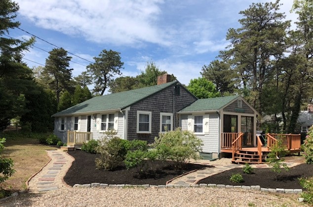 A charming cottage is framed by a wooded landscape, featuring a shingled exterior and a green roof. A pathway made of stones leads to the entrance, bordered by low shrubs and greenery. A wooden deck extends from the side, inviting relaxation in the outdoor setting.