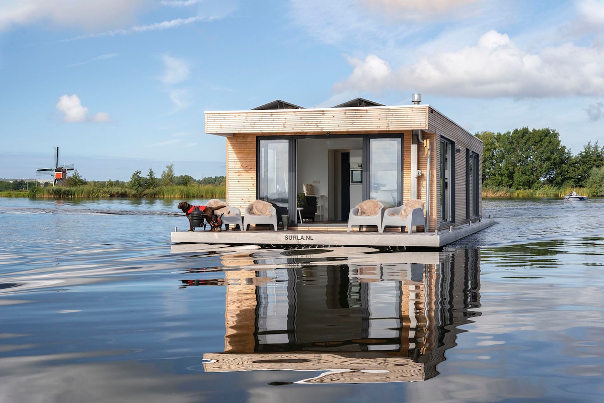 A contemporary houseboat features a wooden exterior and large glass doors that provide unobstructed views of the surrounding water. The serene surface reflects the structure, creating a harmonious connection with the natural environment.