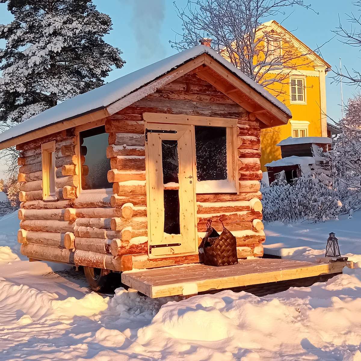 A charming log cabin is set against a snowy landscape, showcasing a rustic exterior with large windows. The entrance features a wooden door and a small porch. Snow covers the ground, and a warm glow emanates from a nearby building highlighted in a contrasting yellow.