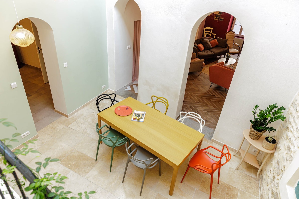 A spacious dining area features a large wooden table surrounded by colorful chairs in various styles. Soft, natural light floods the room through arched openings, which lead to a cozy living area. The floor is finished with light-colored tiles that enhance the welcoming atmosphere.