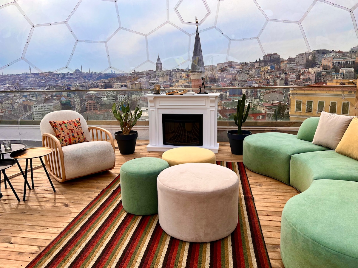 A stylish rooftop lounge area is showcased, featuring a cozy dome structure with a panoramic view of the city. A large sectional sofa in soft green hues, circular ottomans, and a striped rug complement the space. Potted plants and a modern fireplace enhance the inviting atmosphere.