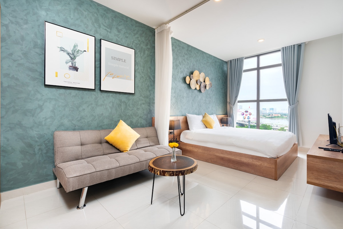 A modern living space features a queen bed and a comfortable sofa, complemented by a decorative wall with artwork. Large windows allow natural light to fill the room, while a small table with a vase adds a touch of warmth to the area.