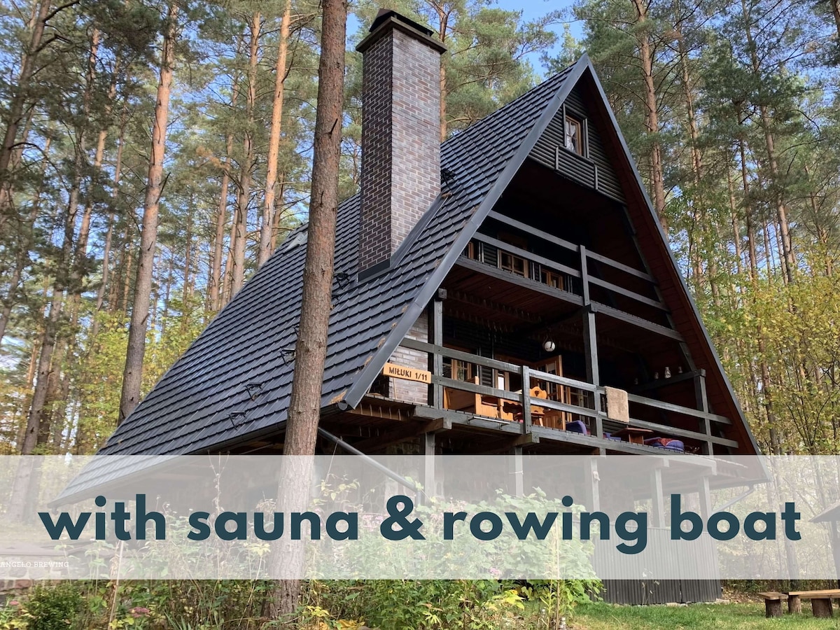 The A-frame house, surrounded by tall trees, features a dark, sloped roof and large wooden balconies. A chimney is visible, suggesting warmth inside. The landscape includes lush greenery, enhancing the natural setting, and a rowing boat can be seen nearby, adding to the outdoor ambiance.