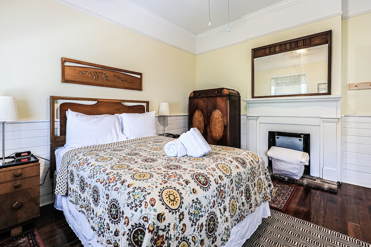 A cozy bedroom showcases a spacious bed adorned with a colorful quilt. A vintage chest of drawers and a large mirror enhance the room's charm. Natural light filters in through the window, and a decorative fireplace adds a historic touch.