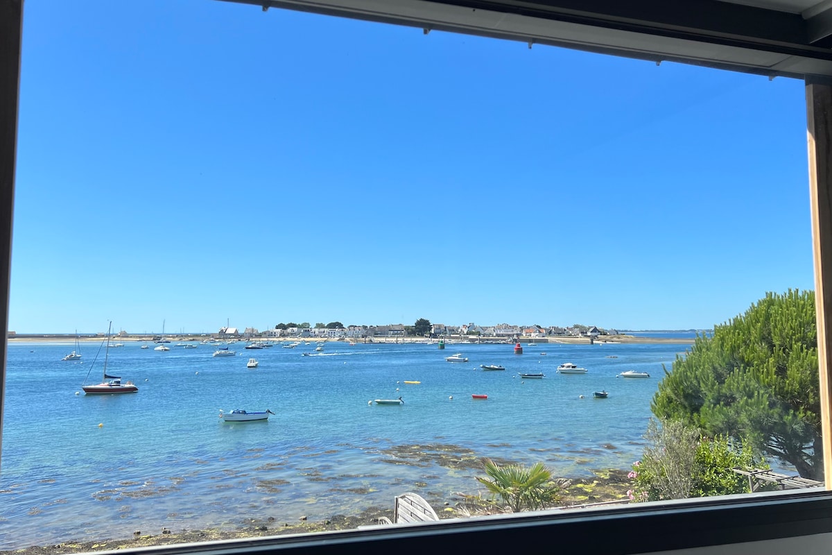 A scenic view of the bay is visible through a large window, showcasing clear blue waters dotted with boats. The coastal landscape features charming buildings in the distance and lush greenery along the shoreline, enhancing the tranquil coastal atmosphere.