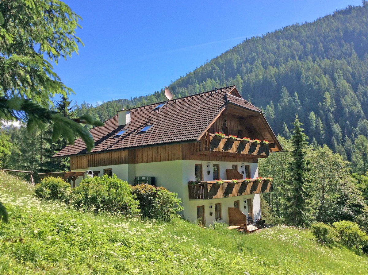 A charming two-story building is nestled in a lush green landscape. Multiple balconies adorned with flower boxes overlook the surrounding trees and hills. The wooden facade and bright white walls contribute to the appealing mountain retreat atmosphere.